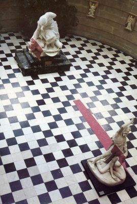 Photo looking down into a rounded room with a black and white tiled floor, containing two large figurative sculptures. A red streak of plasticine partially covers both sculptures and a section of the floor.