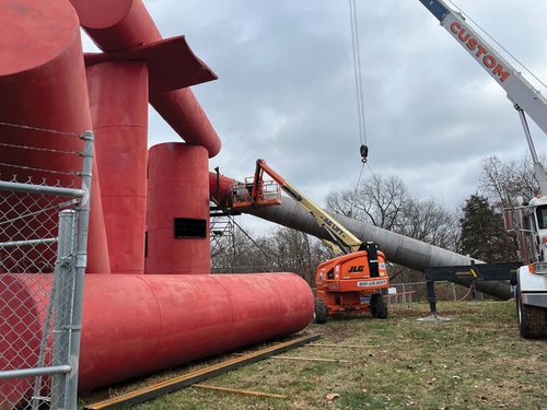 A crane, boom lift and scaffolding surrounding a large, badly-weathered outdoor sculpture made up of several metal cylinders.