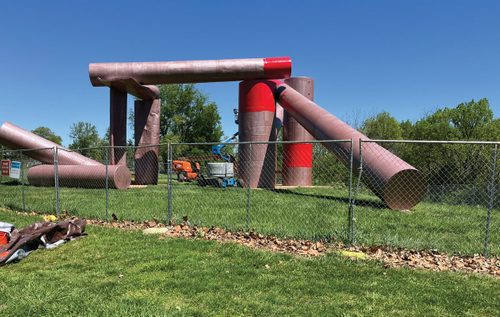 A large, badly-weathered piece of outdoor sculpture, made from metal cylinders piled together in a semi-haphazard way, behind a chain-link fence. Part of the sculpture has been repainted in bright red; the rest is very dull, having been stripped of paint.