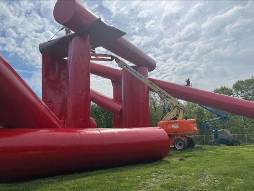 Two boom lifts enabling workmen to paint a large outdoor sculpture made up of several metal cylinders.