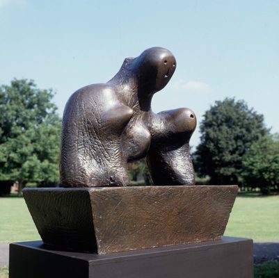 A bronze semi-abstract sculpture with shapes resembling two faces and a breast, sat on a bronze plinth surrounded by grass and trees.