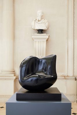 An abstract sculpture carved from black stone sits on a plinth in front of a white portrait bust.