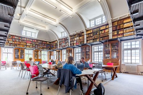 A large, bright library reading room, with two rows of large wooden tables in the centre. Several people are sat there working on laptops.