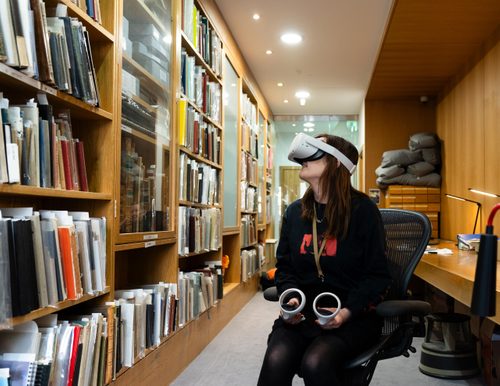A young woman wearing a VR headset, sat on an office chair in an archive.