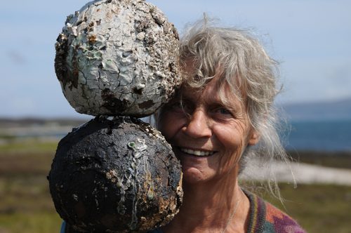 Portrait photo of a woman with grey hair. She is standing with her face partially obscured behind a sculpture, comprised of two head-sized ceramic objects, the top one painted white and the one underneath black.