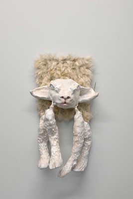 A ceramic head of a sheep, with ceramic sheep's legs hung from its cheeks in the fashion of earrings, attached to a white fleece on a white wall.