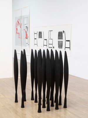 Around 20 identical tall, slender black sculptures are clustered together on a wooden gallery floor. Behind them, some abstract prints are hung on the white wall.