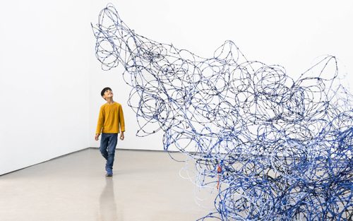 A young boy wearing yellow walks through Lungiswa Gqunta’s sculpture 'Ntabamanzi', made from barbed wire and blue wool. The sculpture is designed to look like a wave breaking against the wall of the gallery.