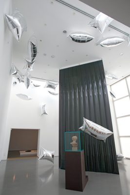 A display cabinet with the head of a damaged marble statue, surrounded by floating, pillow-shaped silver balloons.