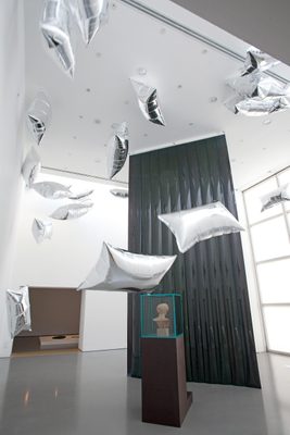 A display cabinet with the head of a damaged marble statue, surrounded by floating, pillow-shaped silver balloons.
