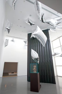 A display cabinet with the head of a damaged marble statue, surrounded by floating, pillow-shaped silver balloons.