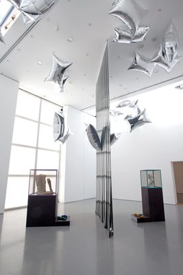 Two display cabinets holding damaged marble statues, separated by a vertical plastic sheet, and surrounded by floating, pillow-shaped silver balloons.