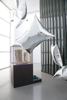 A display cabinet with the lower legs of a broken marble statue, surrounded by floating, pillow-shaped silver balloons.
