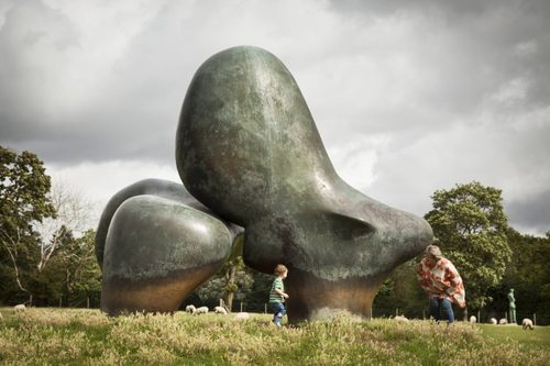 An older woman and toddler playing peek a boo around the base of a bronze sculpture which is standing in a sheep field