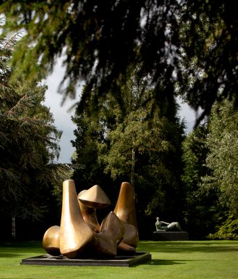 A large abstract bronze sculpture is in the foreground, with a second figurative bronze sculpture in the distance behind, sited on a lawn with trees surrounding.