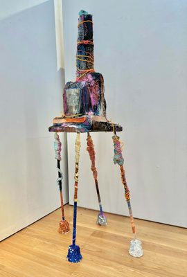 Sculpture of a mill or similar building with a tall chimney, displayed on stilt-like legs. It is colourfully painted and wrapped around with coloured strings.
