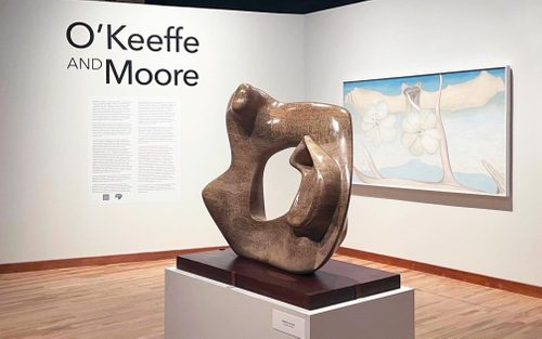 An abstract sculpture by Henry Moore, carved from stone with a void in the centre. Behind this is a painting by Georgia O'Keeffe of flowers, antlers and bones, done in soft shades of cream, white and blue. Text on the wall reads 