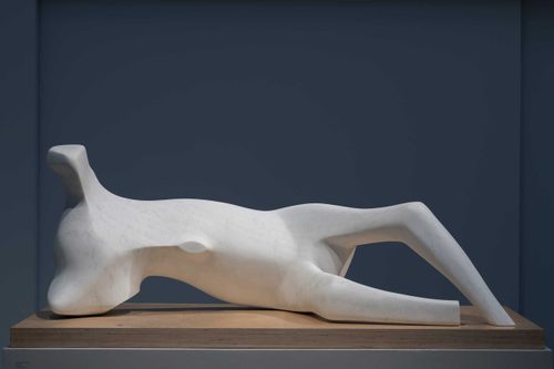 Sculpture of an abstract human figure in a reclining pose, carved from white marble by Henry Moore. The sculpture notably lacks any arms, feet or a head.