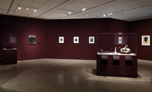 Wide shot of a gallery space, with several small paintings hung on a burgundy wall, and four small sculptures in two display cases.