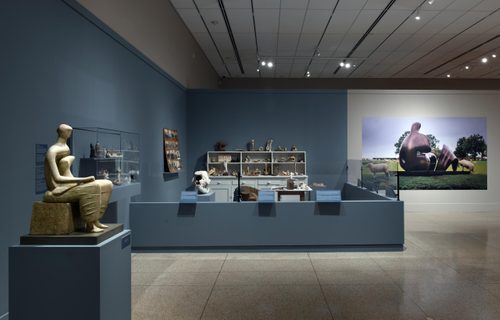 Wide shot of a gallery space containing a recreation of Henry Moore's studio, alongside a stone carving of a seated woman holding a child, and a large-scale photo of a bronze sculpture in a field, surrounded by sheep.