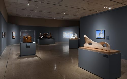 Wide shot of a gallery with blue walls, with four large figurative sculptures in bronze and stone by Henry Moore displayed on plinths. Paintings are hung on the walls further back.