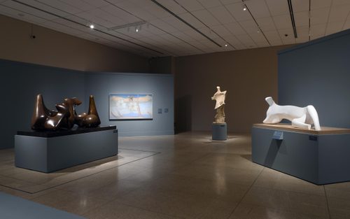 Wide shot of a gallery with blue walls, with three large figurative sculptures in bronze and stone by Henry Moore displayed on plinths, and a painting by Georgia O'Keefe hung on a wall between them.