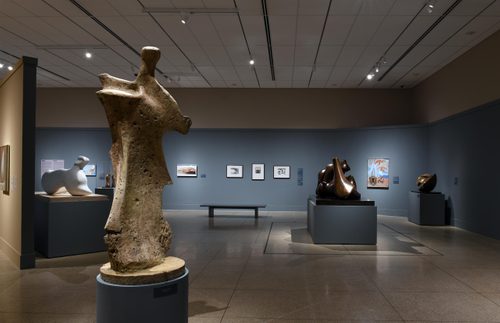 Wide shot of a gallery with blue walls, with a tall sculpture by Henry Moore in the foreground, which looks to be inspired in equal parts by the human figure and pieces of bone. Behind it are more sculptures on plinths, and paintings by Georgia O'Keefe hung on the walls.