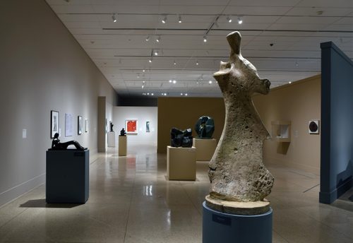 Wide shot of a gallery with sand-coloured walls, with a tall sculpture by Henry Moore in the foreground, which looks to be inspired in equal parts by the human figure and pieces of bone. Behind it are more sculptures on plinths, and paintings by Georgia O'Keefe hung on the walls.