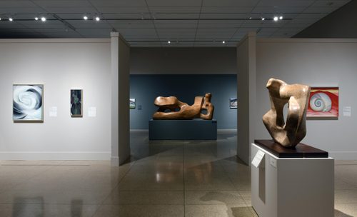 Wide shot of a gallery, with three paintings and an abstract stone sculpture in the foreground, and a wooden carving of a reclining figure visible through a corridor into the room behind.