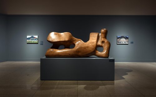A large, wooden carving of a reclining figure by Henry Moore, displayed on a plinth. Two small paintings by Georgia O'Keefe are hung on the blue wall behind, to either side of the sculpture.