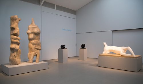 A gallery space filled with sculpture. To the left are two abstract, organic standing figures carved from grey stone. To the right is an abstract reclining figure in white stone.