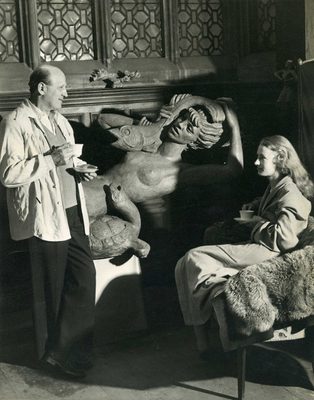 Black and white photo of an older male sculptor having a cup of tea with a young female model, with a sculpture of a mermaid behind them.