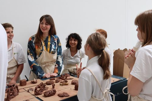 A group of school children taking part in a sculpting workshop, they are listening to the workshop leader and the table in front of them is littered with clay objects in progress