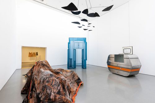 A large gallery space filled with sculptures: Andrea Zittel’s 'A-Z Escape Vehicle, owned and customised by Bob Shiffler' 1996, a silver construction with an orange stripe that looks like a small caravan or trailer; Do Ho Suh’s 'Hub, Wielandstr. 18, 12159 Berlin' 2015, a small, semi-transparent room made from polyester fabric over a stainless steel frame, which visitors can walk inside; Liz Ensz’s 'Convexity / Concavity' 2015, made from fabric piled over objects to resemble a mountain range; and Alexander Clader’s ‘Chicago Black’ 1949, a mobile handing from the ceiling made from large, flat triangular shapes.