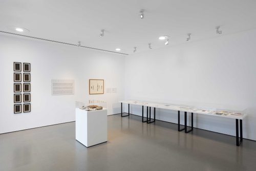 A white-walled gallery space, with frames mounted on the walls and display cases filled with tools, notes and photos.
