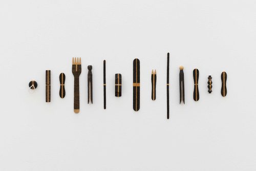 Fourteen everyday wooden objects, including a plug, chip fork, lolly stick, and a peg, are arranged in a line on a wall. They have all been 'decorated' with different patterns of black lines, burnt by the sun using a magnifying glass.