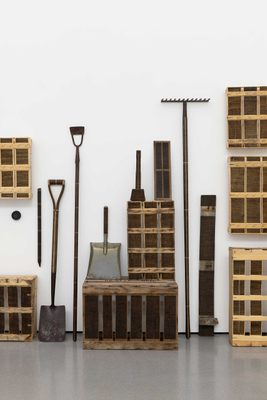 Many of Roger Ackling's wooden works stacked neatly against a wall, including crates, and various tools: a spade, hoe, dustpan, hammer, and rake. They are all decorated with burned lines.