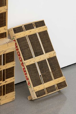 A wooded crate, leaning against a white gallery wall. It has been marked with horizontal stripes, burned into the wood using a magnifying glass and the sun.