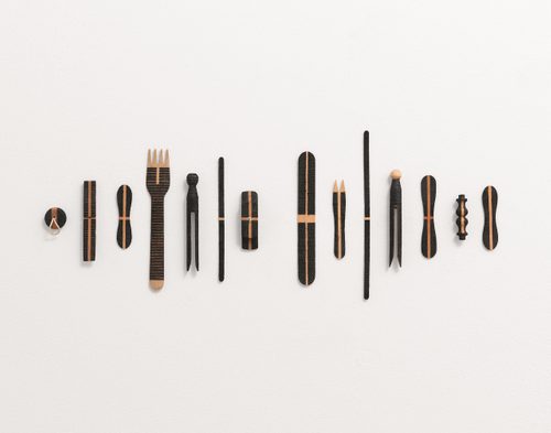 Fourteen everyday wooden objects, including a plug, chip fork, lolly stick, peg are arranged in a line on a wall. They have all been 'decorated' with different patterns of black lines, burnt by the sun using a magnifying glass.