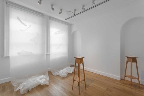 Gallery space with long, translucent paper artworks hung in front of a window, trailing like a roller blind to pool on the floor. There are also two slender wooden side-tables being used as plinths to display tiny 'ice watch' sculptures'.