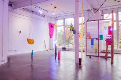 Art installation with many colourful plastic objects scattered throughout a gallery and suspended on coloured string from the ceiling.