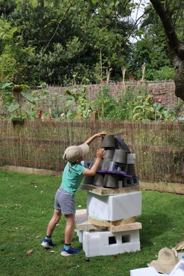 A young child wearing a baseball cap stretches up to balance a plant pot on top of a sculpture made from packing foam and plant pots. He is working in a garden, with a fence and vegetable plants behind him.