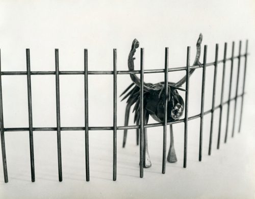 A small, cartoonish steel sculpture of a moose or stag behind a wire fence, poking its nose through a gap.