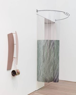 A photo showing a contemporary sculpture installed in a gallery. Two abstract forms, in pink and verdigris hang from a white wall.