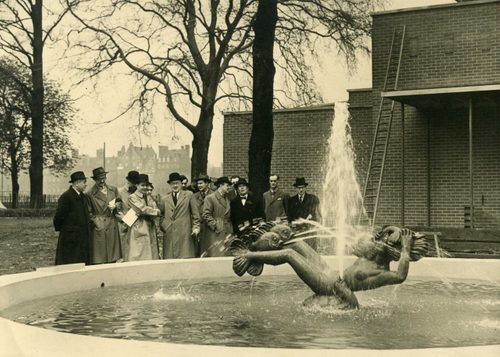 Black and white photo of a fountain-sculpture depicting a mermaid, turtle and fish spraying water. A group of formally dressed men wearing long coats and hats are stood behind the fountain.