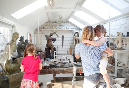 A woman and two young girls, with their backs to the camera, look at Henry Moore’s Top Studio. The space is laid out as Henry Moore would have used it in his lifetime, with various wood, plaster and stone carvings dotted around the room, and numerous tools out on top of workbenches or hung on the walls.