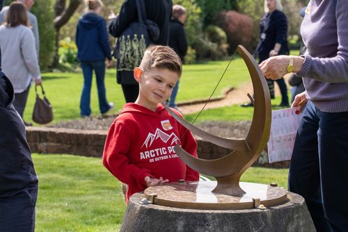 A young boy in a red sweatshirt looks curiously at a small bronze sundial