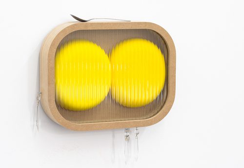 A colour photo showing a contemporary sculpture. The piece is suspended on a white wall. It is an oval mdf cabinet, which contains two lemon yellow circular forms. Resting on the top edge of the cabinet is a spoon. Suspended from the bottom are crystal bed drops, like a chandelier