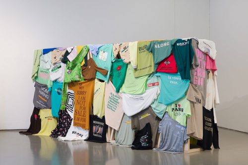 Photo of Shanzhai Lyric's sculpture 'Incomplete Poem (hedge)'. The made from numerous colourful t-shirts arranged on a wooden frame, and are part of a larger collection of bootleg slogan shirts, often featuring mistranslations. Some of the slogans on these t-shirts read: 'SIMPLE TRUTH INSIDE', 'NOM HIRING', 'NUIVERSAL', 'I AM NOT SORRY I AM NOT FOR SALE I AM FOR I AM NOT FOR SALE', 'BEWARE OF PEOPLE LIKE ME', 'Be So Rooted In Yourself That Naomhetu Absence Or Presence Can Disturb Your Inner Peace', 'MISSONE fashion', 'ALONG', 'BRAVE', and 'la vieen rose'.