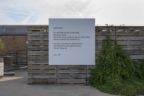 A square board with a poem on it, attached to a wooden wall outdoors. The poem reads: 